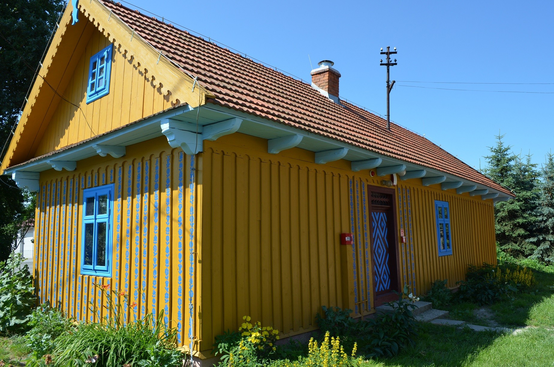 Drewniana chałupa pomalowana na żółto, z niebieskimi ramami okien.